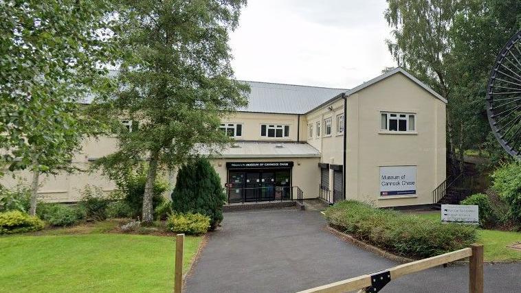 A cream-coloured building with a large, black-painted miner's wheel to the side of it. The miner's wheel appears taller than the two-storey building. There is a tarmac driveway and gate in front of an entrance which has a ramp and glass doors. There are trees surrounding the building and a lawn in front.