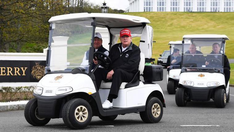 Donald Trump and a passenger in a golf buggy next to a black and gold sign which says "Trump Turnberry". Trump is wearing a red cap which reads "make America great again" and black clothing, he is wearing white golf shoes.