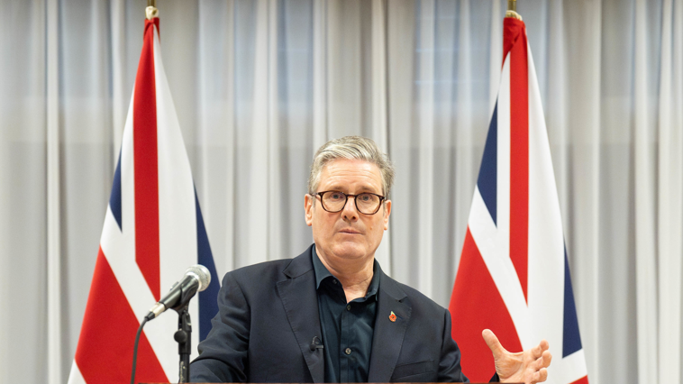 UK Prime Minister Sir Keir Starmer in dark shirt and dark suits stands in frnt of two Union Jack flags to give speech