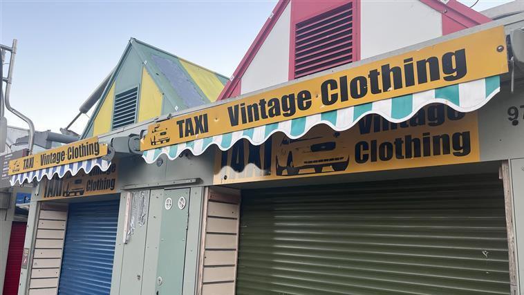 A stall on Norwich market with green shutters. A yellow sign reads, TAXI VINTAGE CLOTHING.