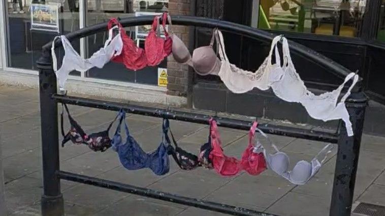 Different coloured bras have been hung on railings in a street with shops. 