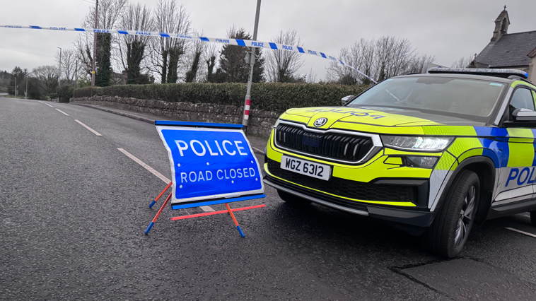 A police SUV next to a sign saying "police, road closed" and cordon police tape blocking the road. there is a stone wall, with a hedge running down the road. 