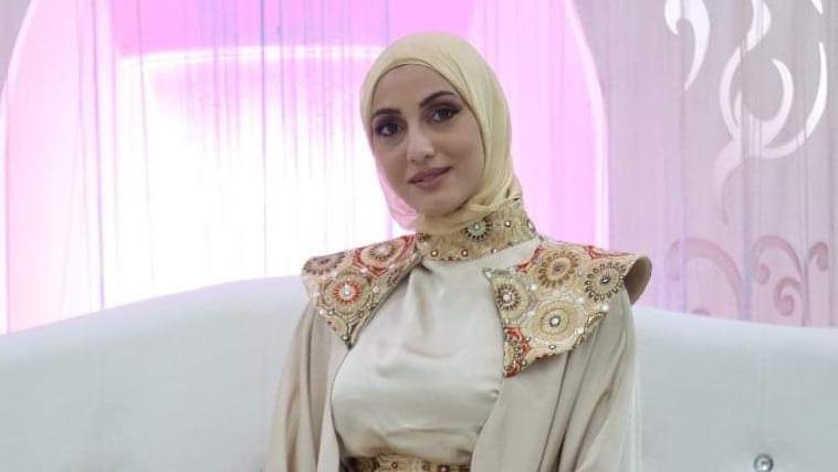 A young woman smiles at the camera. She is wearing a white and ivory dress with an ornate gold patterned collar and shoulder pads. She is sitting on a white couch. Pink light glows in the background.