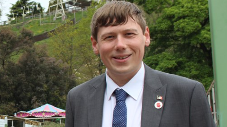 A man with short brown hair and wearing a grey suit, white shirt and tie. He is standing by a grassy hillside with trees and fences and smiling at the camera.