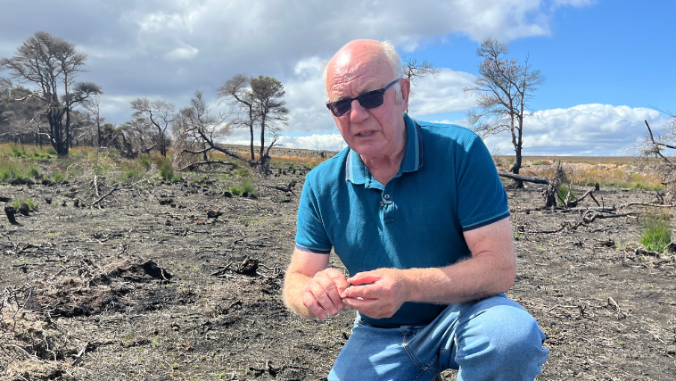 Lol Hodgson is kneeling on blackened moorland. He is wearing dark sunglasses and a blue t shirt and blue jeans