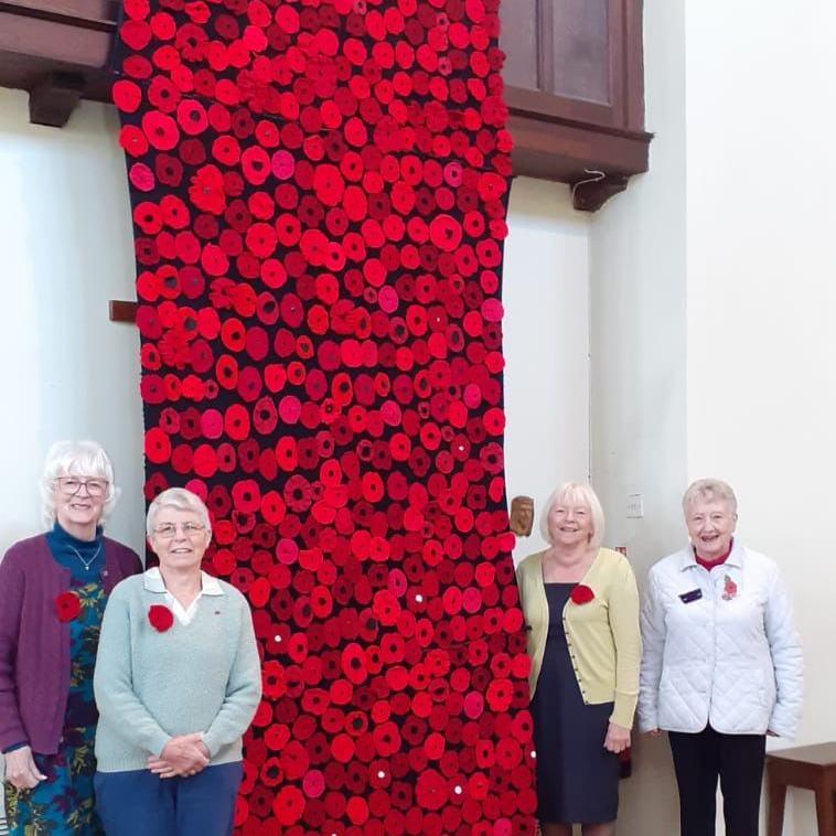 A tapestry of knitted poppies hangs on an inside wall in a church, with two women stood either to it. All of the women are wearing poppies on the left side of their chests and are smiling next to the display.