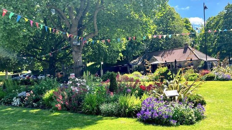 The village green with purple, pink and red flowers. The lawn is freshly cut and there is colourful bunting hanging from a tree. 