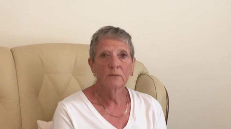 A woman sits on her sofa. She is wearing a white top and had a necklace on. She's sat on a pale sofa.