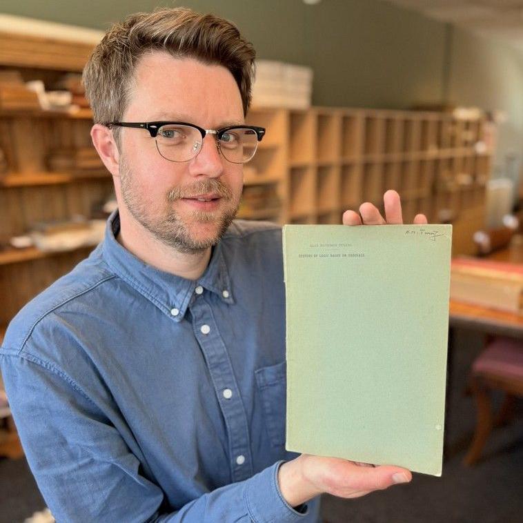 A man with short brown hair, glasses and a blue shirt, carefully holds up an old paper while standing in an archive office.