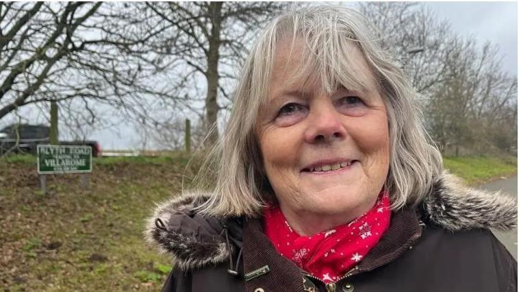 Head and shoulders shot of Penny Carpenter smiling and wearing a brown winter coat with a fur trim and a red scarf. She is standing in a road with a green verge and trees behind.