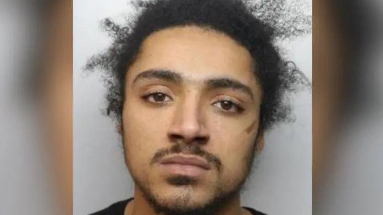 A police mugshot of a man with black hair which is sticking up in places. He has short stubble and is wearing a black T-shirt.