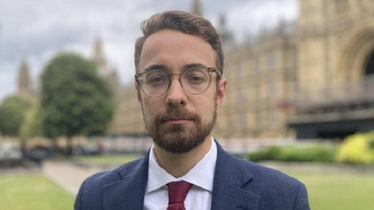 Luke Charters MP standing outside Parliament. He is wearing a navy suit and a maroon tie. He has glasses, brown hair and a beard and is looking at the camera with a serious expression. The Houses of Parliament can be seen in the background.