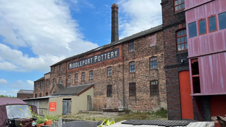 The brown brick building has a large Middleport Pottery sign in black and white high up. A chimney can be seen behind and a red painted door and a pink building can be seen on the right of the image at ground level. The building appears to be by water with a small maroon boat moored a few feet away.