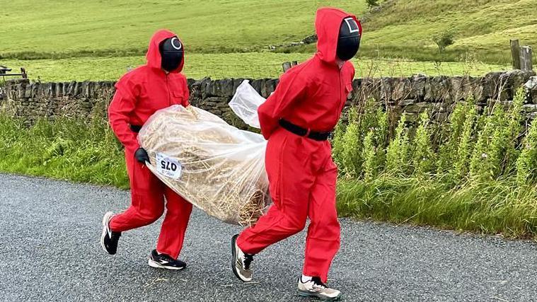 Two people in red full-body jumpsuits, black face masks and black belts are carrying a bale of hay between them.