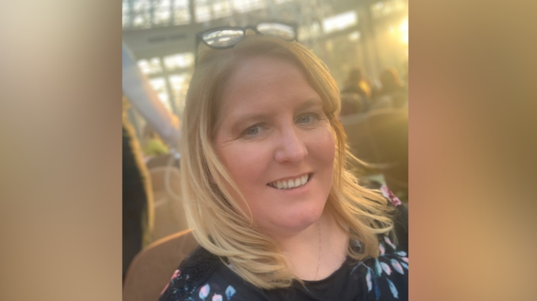 Ciara McCay. She has shoulder-length blonde hair, wearing glasses on her head and a black floral top.