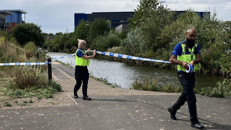 Two police officers unroll police tape at the scene, one is male with a black beard and the other female with blonde hair. They have attached the tape to a post next to a path by the canal