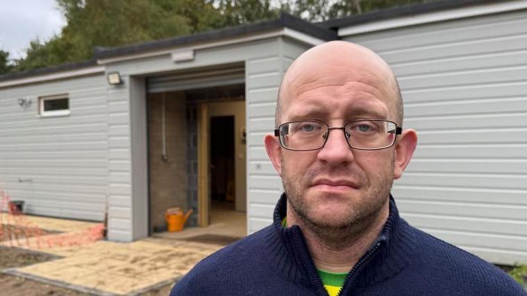 Peter Clarke has a shaved head and close cropped beard and moustache. He is wearing glasses, and is wearing a yellow teeshirt with a green trim collar, and a blue cardigan over. He is standing outside the Belton Scout Hut extension, which is incomplete in build.