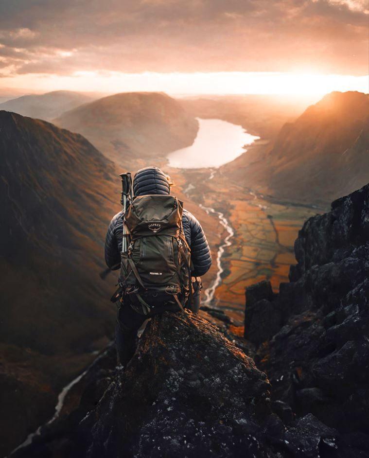 A hiker in a puffer jacket with a large backpack sits on a rock with their back to the camera, looking out across a valley and lake with the sun coming through the clouds.