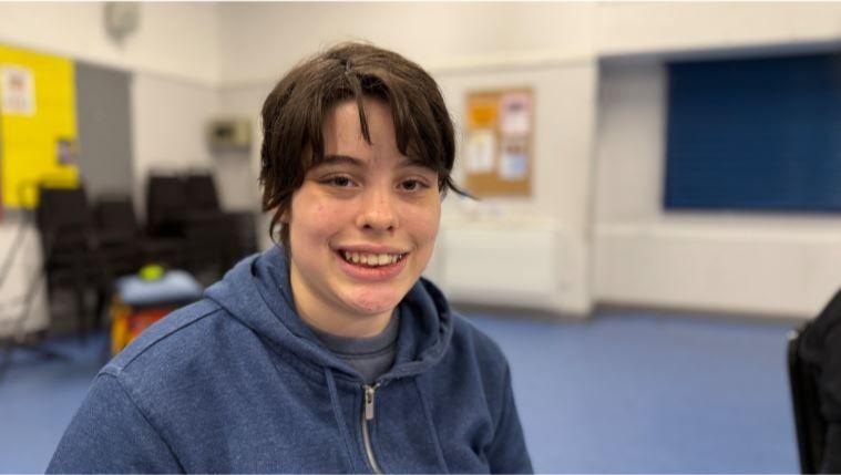 Grace is sitting down and smiling at the camera. She has dark short hair and a fringe and is wearing a zip-up navy hoodie.