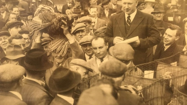 A sepia picture of a group of men in suits, caps and hats selling birds near cages. One man holds a turkey above his head