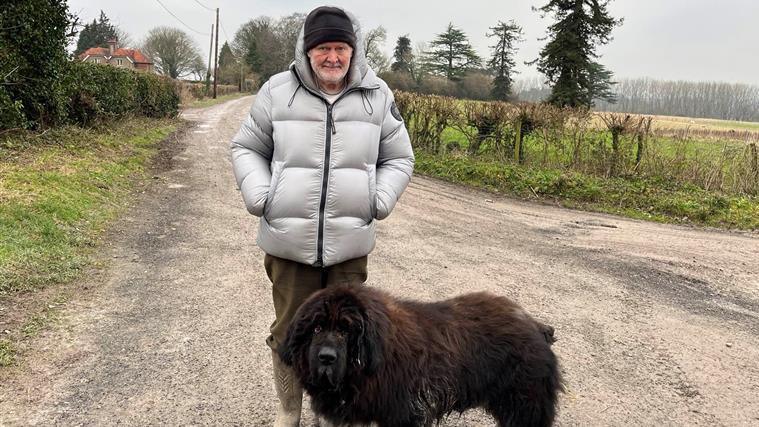 A man with white stubble, a black beanie, a silver coat and green trousers and wellies looking at the camera while standing in front of a large, black, hairy dog. They are standing on a country road.