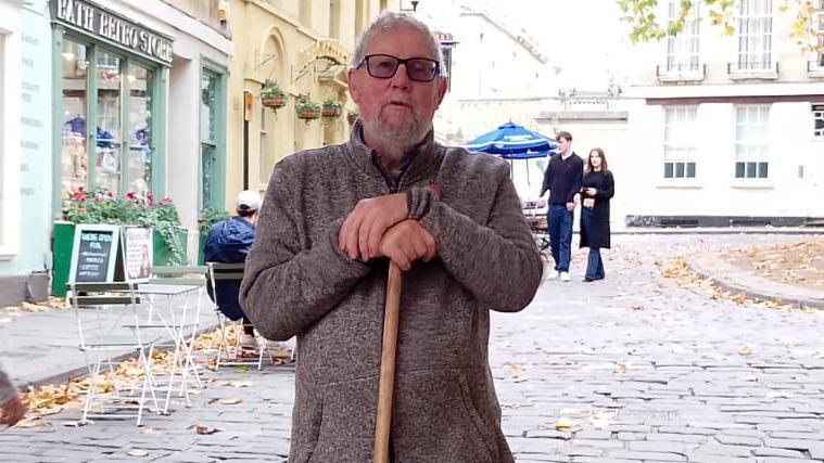A man stands in the centre of a cobbled street. He is wearing a thick brown jumper and is leaning on a brown stick. He has grey hair and glasses and behind him are a man and woman walking. There is also a shop with a sign and tables and chairs. 