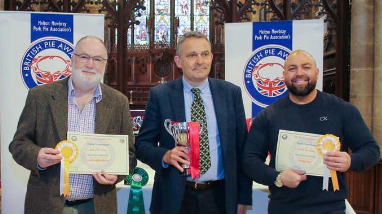 Alex Melling from Baldy's Dark Kitchen stands holding a certificate to the right of a competition judge and another competition winner at the British Pie Awards. 