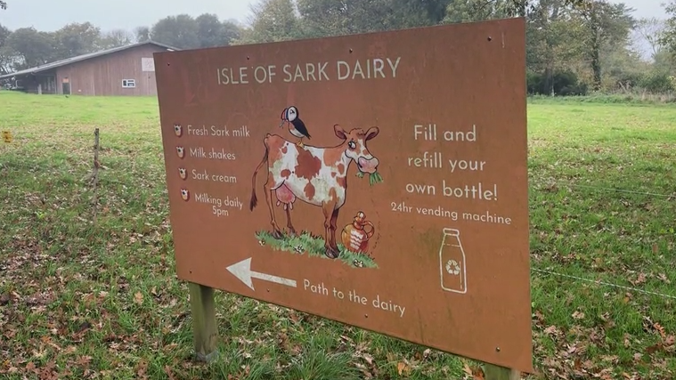 A rustic large sign stands in a grassy field near a wooden with what looks to be a slate roof, surrounded by leafy trees. The sign reads “Isle of Sark Dairy” in bold letters and features playful illustrations of a black-and-white cow with a puffin perched on its back, a glass milk bottle, and a traditional metal milk churn. Below the title, the sign lists offerings: fresh Sark milk, milkshakes, Sark cream, and notes that milking occurs daily at 5pm. It encourages visitors to fill or refill their own bottles using a 24-hour vending machine. An arrow points left with the words “Path to the dairy.”