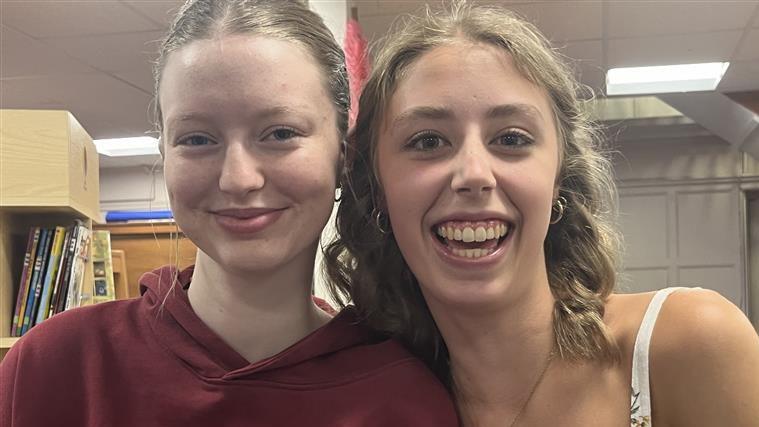 Two young women, one with her hair pulled back into a right ponytail and the other with shoulder-length blonde curled hair, smile at the camera