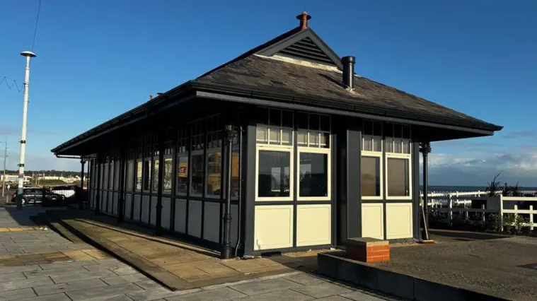 A former tram shelter which is now a cafe on Seaburn's seafront. It is a single-storey building with white panelling on its lower section with windows above. The detailing between the windows and panels is in black, while a dark tiled and pointed roof sits on top. The sea appears in the distance and to the right of the building.