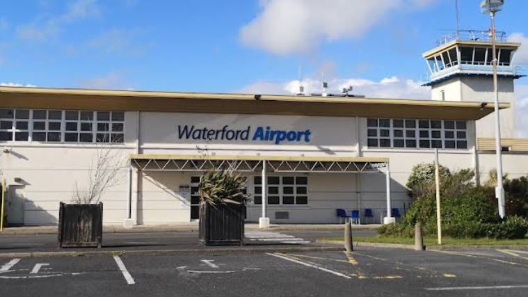 the image shows the exterior of Waterford airport in the Republic of Ireland. It is a white building with a high tower on the right of the picture. The words 'waterford airport' are written in blue above the entrance