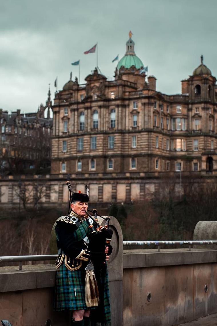 A bagpiper wearing a green kilt standing on a bridge in Edinburgh while playing his instrument. A grand looking building is in the background.