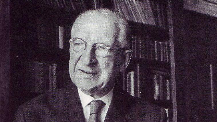 A black and white image of Eamon de Valera, with metal round glasses and short grey hair. He is wearing a suit jacket, shirt and tie. Behind him is a bookcase.