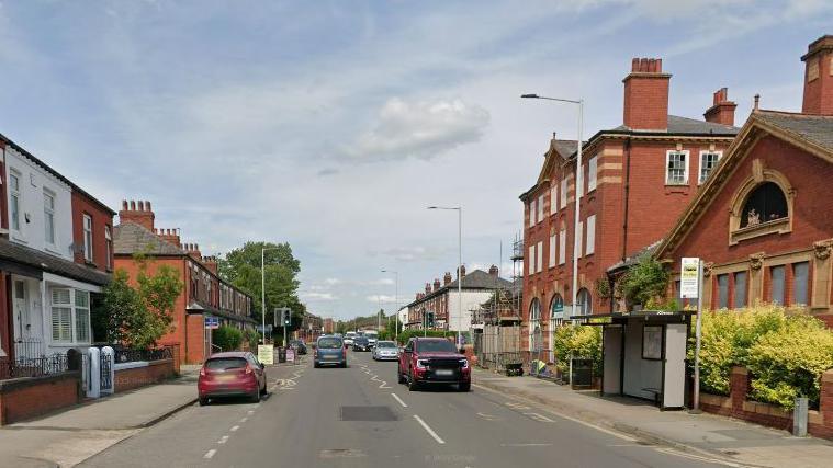 Google streetview of cars driving along Gorton Road past a bus shelter with houses and a civic centre on either side.