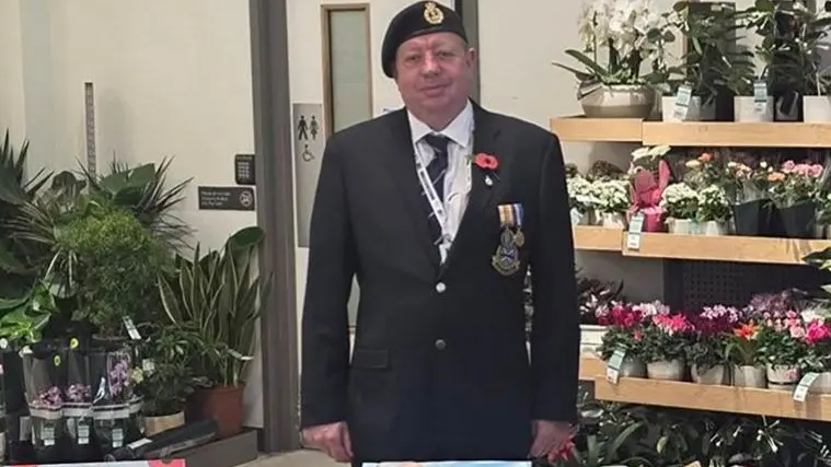 Andrew Johnson is stood in the middle of the image looking towards the camera. He is stood behind a table which has poppy's on for an appeal. He is wearing a forces suit and is stood in front of flowers.
