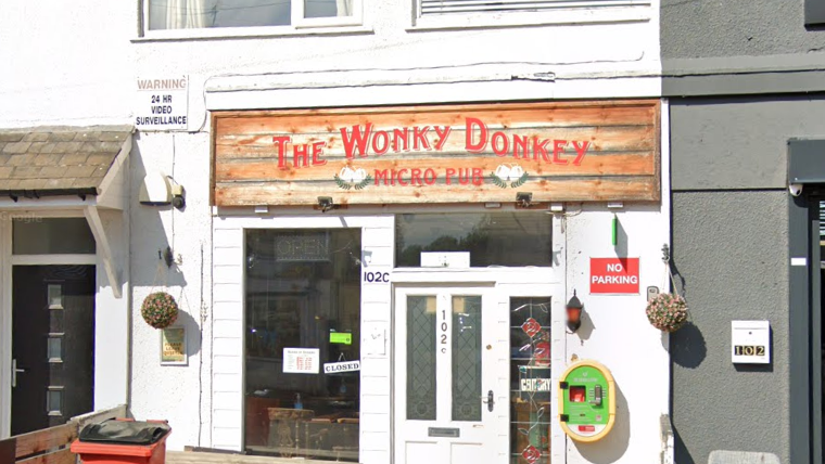 Frontage of the pub building- with a white door - a red bin in front and a green and yellow defibrillator on the side of the building 