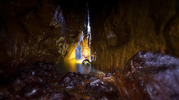 A man wearing safety gear is up to his chest in water in an abandoned mineshaft . His head torch lights up the rocks in front of him