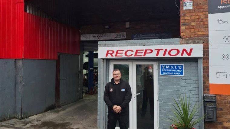 A man standing by a door at the reception of a garage. He is wearing a black uniform and the door has a sign on it saying "MOT reception and waiting area." There is also a large sign in red above him saying "Reception" and a plant with long green leaves. 