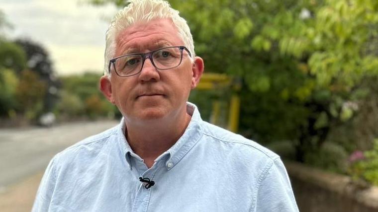 Terry Cunningham, with white hair and black-framed glasses, wears a pale blue shirt. He is standing by a lilac wreath tied to a lamppost at the spot where his mother was fatally struck by a van.