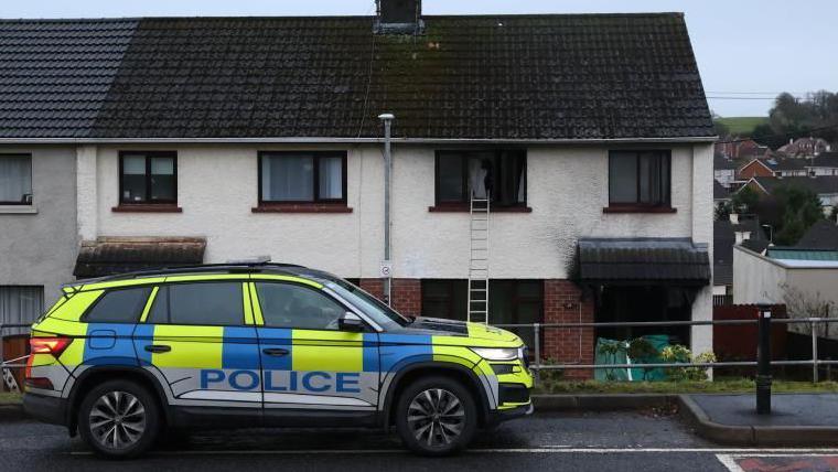 A yellow and blue police car is parked outside a row of terrace houses. The house on the right has burnt out windows and a ladder leading to the upstairs left window.