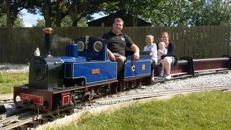 A small blue and black steam train with "C R" on its side and a red front plate labeled "CR 273" sits on a narrow-gauge track. Four people are riding the train: an adult man in front, two children in the middle, and a woman holding a baby at the back. The background shows green grass, trees, and a wooden fence in a park-like setting