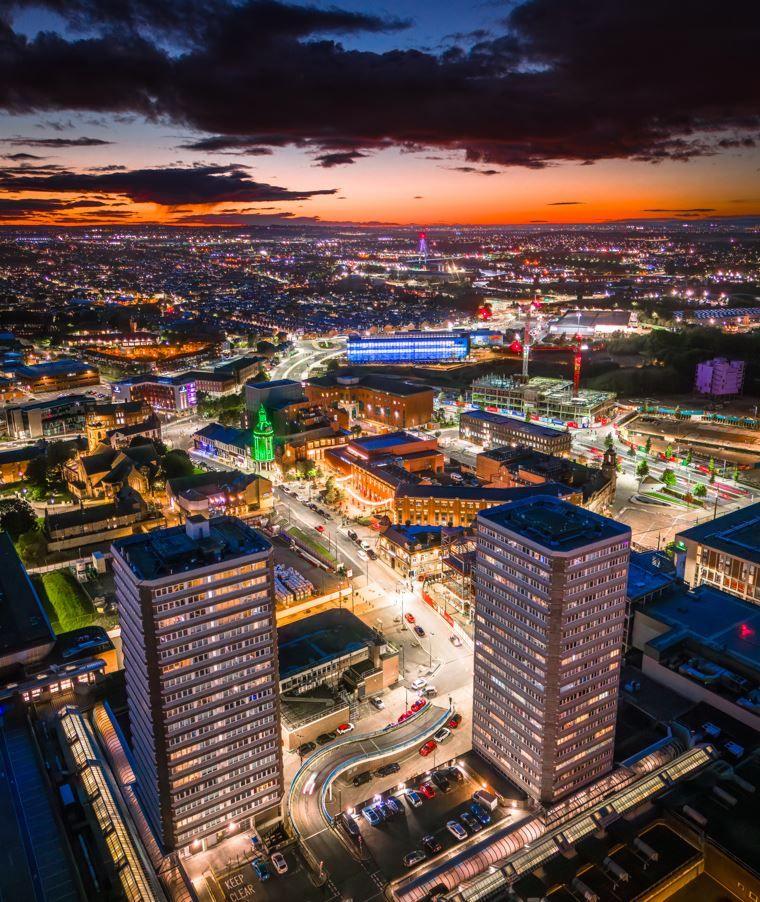 Aerial view of two tower blocks and lit city streets, with an orange sky and dark clouds in the background.