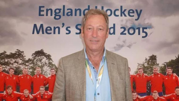 An archive image of the late Graham Wilson. He is smiling for the camera in front of a poster of a hockey tea, that reads England Hockey Men's Squad 2015. Their kit is red.