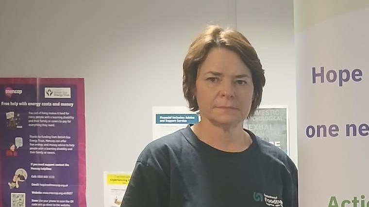 Diane Kindred, with short brown hair, wearing a black top, stands in a room with public information posters.