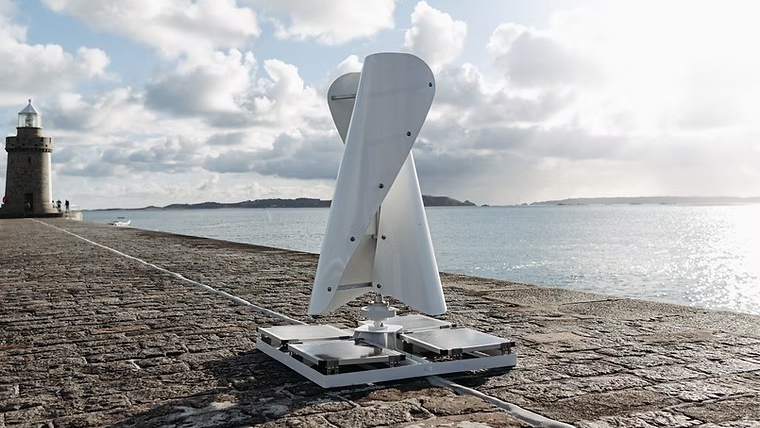 A white wind turbine on a granite pier with a lighthouse and some islands in the background. The wind turbine is made of two curved, vertical sails on a flat base with solar panels on it. It is 1.8m (5.9ft) tall.