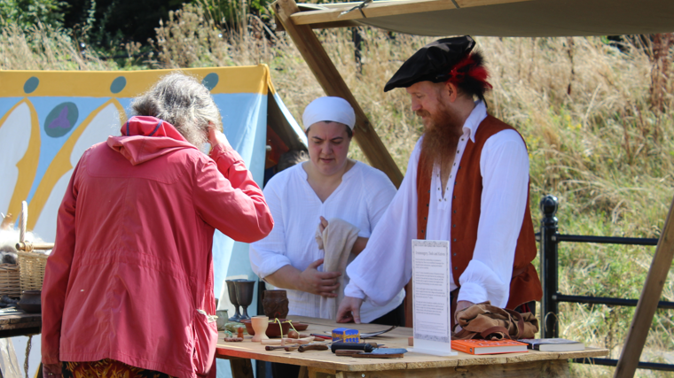 Two stall holders are selling items at the outdoor fair while wearing traditional dress. The items on the table appear old fashioned. The stall holders - a man and a woman - are talking to the customer as she browses the items on the table.