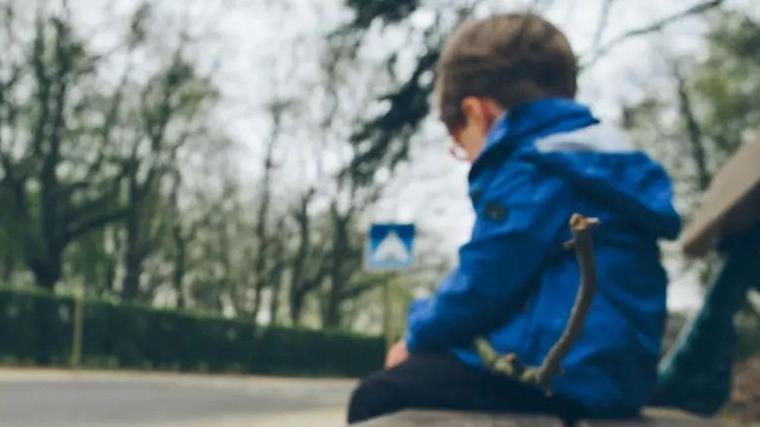 A lonely-looking child sitting on a bench, wearing a blue coat. He has short, dark hair and is wearing glasses while staring down at the pavement.  There are trees, general foliage and a bush on both sides of the road that runs past the bench.