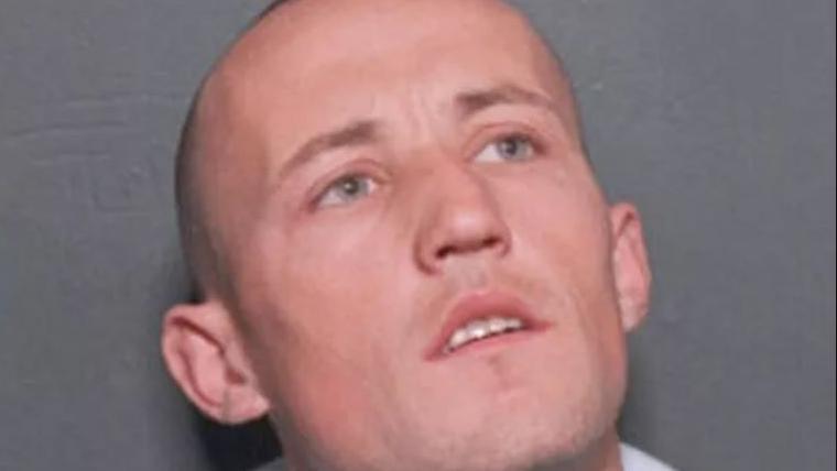 Police mugshot of a shaven-headed Kevin "Gerbil" Carroll. His mouth is open but he is not looking at the camera. Instead his head is slightly tilted up and his blue eyes are fixed on a spot behind the camera on his left. 