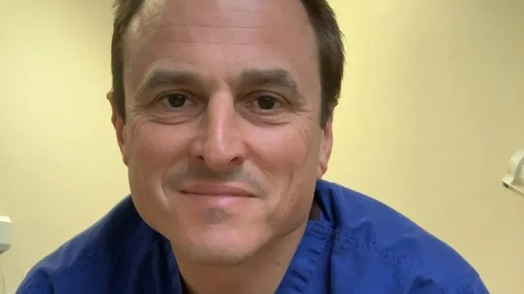 Paul Williams, who has brown hair and is wearing blue scrubs, smiles into the camera.