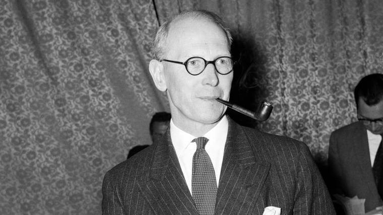 Sir John Wolfenden, who is smoking a pipe, wearing a dark suit.